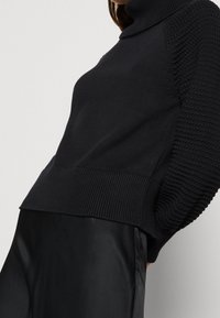 Schwarzer gestrickter Rollkragenpullover mit gerippten Besätzen und strukturierten Ärmeln, kombiniert mit einem eleganten schwarzen Rock unterhalb der Taille.