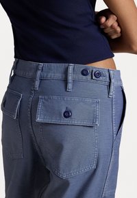 Pantalon en denim bleu clair avec fermeture à double bouton, présentant deux poches arrière avec rabat et accents de bouton. Texture lisse et coupe ajustée.