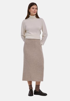 Femme debout les mains derrière le dos, portant un pull à col roulé crème, une jupe midi en maille beige, des chaussettes marron et des mocassins noirs.