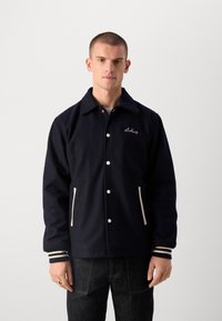 Jaka no tumši zila vilnas maisījuma ar pogu aizdari, divām priekšējām kabatām un baltām svītrām uz piedurknēm. Iekļauta izšūšana uz krūšu.