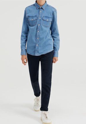 Person, der ein hellblaues Jeanshemd mit Knöpfen, dunkelblaue Hosen und weiße Sneaker trägt, steht vor einem weißen Hintergrund.