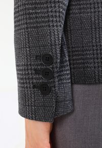 Manche d'un blazer à motif pied-de-poule gris foncé avec trois boutons noirs au-dessus du poignet d'une personne et une partie d'un pantalon gris.