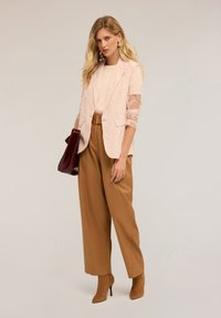 Hellrosa Spitzenblazer mit transparenten Ärmeln über einem beigen Top, kombiniert mit hochgeschnittenen braunen Hosen und braunen, absatzschuhen. Burgunderfarbene Handtasche.