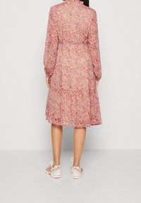Robe florale rose avec de longues manches bouffantes, ourlet en plusieurs couches et tissu texturé. Associée à des chaussures de sport beiges avec des accents rouges.
