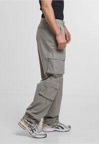 Pantalons cargo gris avec poches latérales, fabriqués en tissu léger. Coupe ample avec des détails de coutures ; associés à des chaussures de sport noires et argentées.