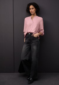 Blusa rosa chiaro con colletto arricciato e bottoni, abbinata a jeans neri a vita alta con gamba larga. Il modello indossa stivaletti neri alla caviglia.
