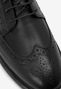 Scarpe eleganti in pelle nera con dettagli in brogue, caratterizzate da motivi perforati e una finitura liscia, stringate, con punta arrotondata e silhouette affusolata.