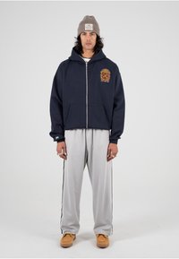 Marineblå zip-up hettegenser med brodert våpenskjold, kombinert med lysegrå joggebukser med sidelisting og brune slip-on sko.