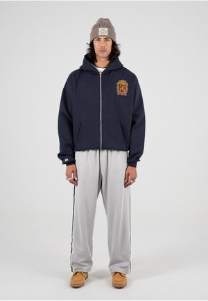 Mørkeblå zip-up hoodie med broderet våbenskjold, parret med lysegrå sweatpants med sidelangs striber og brune slip-on sko.