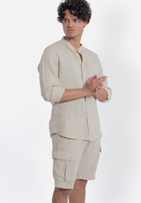 Camicia in lino beige con colletto alto e maniche lunghe, abbinata a pantaloni cargo dello stesso colore con tasche laterali e una vestibilità comoda.