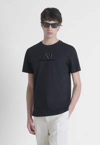 Black cotton t-shirt featuring a textured logo in the center. Round neckline, short sleeves, paired with light-colored pants.