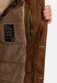 Chaqueta de pana marrón con un forro de borreguito beige, que presenta una etiqueta interior, bolsillos con cremallera y tejido texturizado.