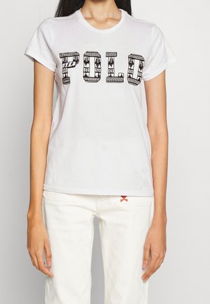 Person trägt ein weißes T-Shirt mit dem Wort "POLO" in schwarzen gemusterten Blockbuchstaben und weiße Hose mit einem roten Detail an der Tasche.