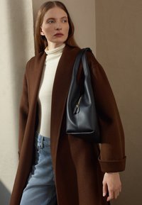 Cappotto di lana marrone sopra un dolcevita bianco, abbinato a jeans azzurri chiari. Borsa di pelle nera con zip dorata e texture morbida.