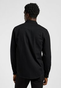 Camisa negra de manga larga hecha de algodón, con un diseño de botón, cuello puntiagudo y dobladillo curvado con una textura suave.