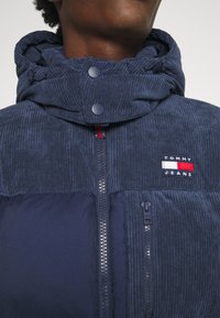 Primo piano di una persona che indossa una giacca in corduroy blu con colletto alto, zip frontali e patch del logo Tommy Jeans.