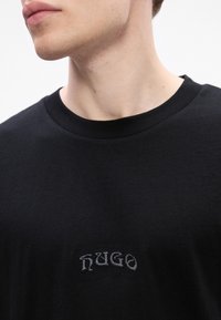 Primer plano de una persona que lleva una camiseta negra de cuello redondo con el pequeño y estilizado "HUGO" bordado en el pecho, mirando ligeramente hacia la izquierda.