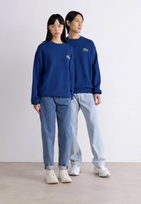Sudaderas azul marino con un pequeño logo bordado, combinadas con jeans de un azul claro y azul oscuro. Ambos llevan zapatillas blancas.