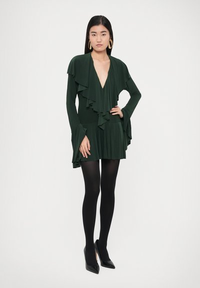 Zielona sukienka z długim rękawem, falbanami, głębokim dekoltem w kształcie litery V oraz rozkloszowanymi rękawami; w połączeniu z czarnymi rajstopami i szpiczastymi szpilkami.