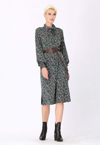Robe à manches longues en tissu vert foncé avec un motif floral, ceinturée à la taille avec une ceinture marron, terminée par des fentes latérales. Bottines incluses.