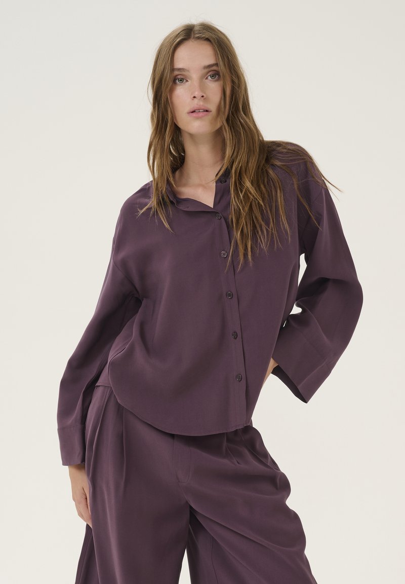 Loose-fitting, button-up shirt in dark purple fabric with long sleeves and a rounded collar, featuring a smooth texture and minimalist design.
