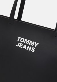 Svart toteväska i syntetiskt material, med vit "TOMMY JEANS" text på framsidan och dubbla kraftiga handtag. Slät yta.