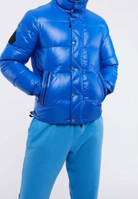 Niebieska pikowana kurtka z wysokim kołnierzem, błyszczącym wykończeniem, zatrzaskami, ukośnym przeszyciem i dwoma kieszeniami na zamek. W zestawie z niebieskimi spodniami dresowymi.