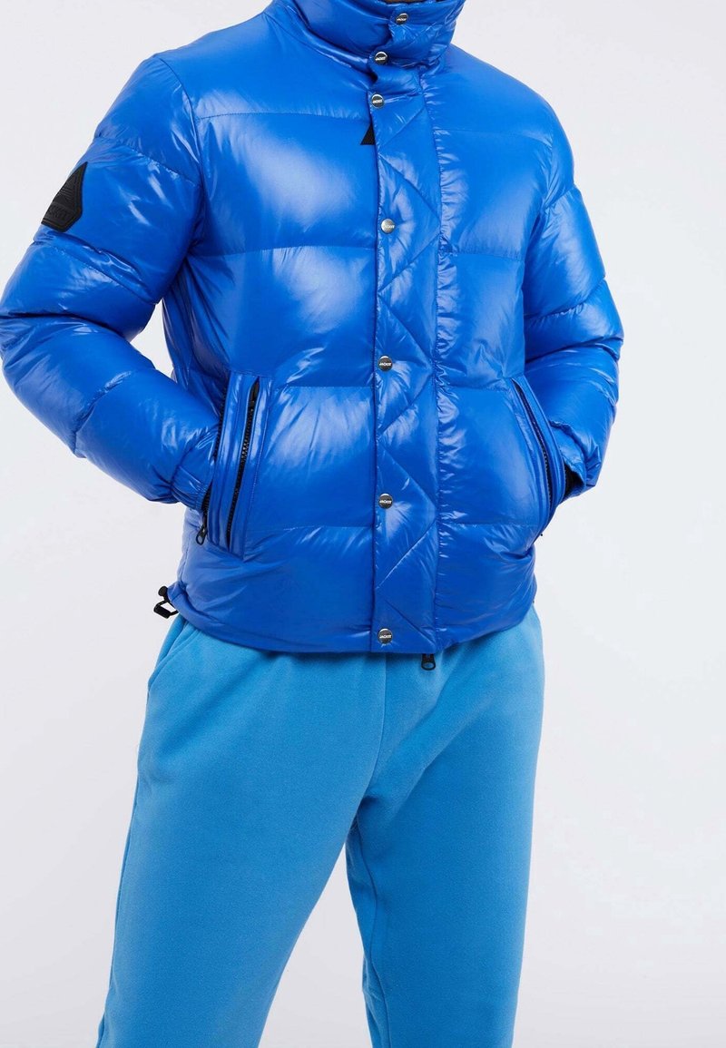 Niebieska pikowana kurtka z wysokim kołnierzem, błyszczącym wykończeniem, zatrzaskami, ukośnym przeszyciem i dwoma kieszeniami na zamek. W zestawie z niebieskimi spodniami dresowymi.