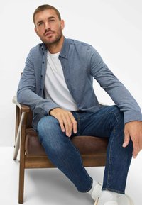 Blaues Hemd mit Knopfleiste und strukturiertem Muster, kombiniert mit dunklen Jeans und weißen Sneakern. Sitzt auf einem braunen Holzhocker.