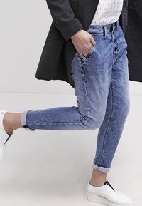 Jean en denim bleu délavé avec une coupe ajustée, dotés de poches latérales et de revers retroussés, associés à des chaussures à plateforme blanches et un manteau sombre.