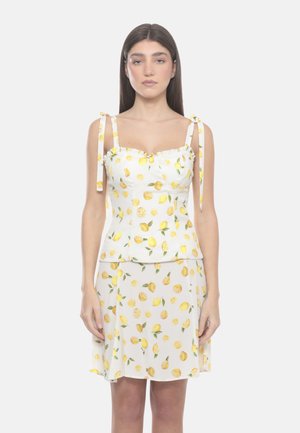 Donna che indossa un vestito bianco senza maniche con stampa di limoni gialli e spalline legate a fiocco, in piedi davanti a uno sfondo semplice.