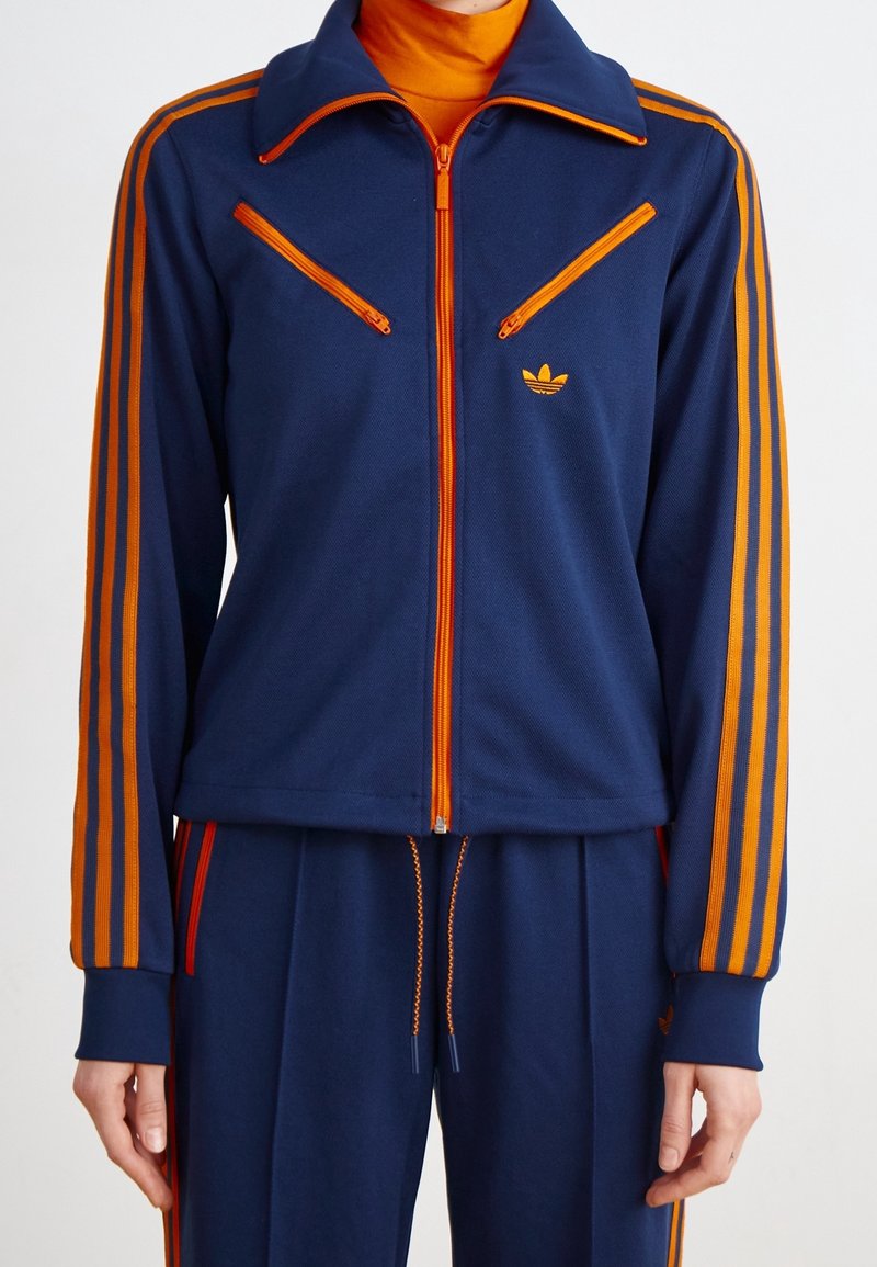 Träningsjacka i marinblått med orangea ränder och dragkedjor. Har en dragkedja vid kragen, två fickor med dragkedja och en broderad logotyp på bröstet.