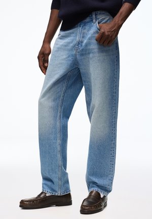 Mand iført lyseblå jeans med lige ben, mørkebrune læderloafers, mørk sweater, stående med den ene hånd i lommen.