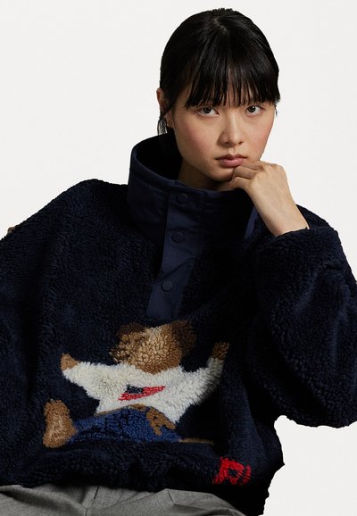 Navyfarbiger Fleecepullover mit hohem Kragen, versehen mit einem Bärenmotiv in Beige und Weiß. Mit Knopfdetails und weichem, strukturiertem Stoff.