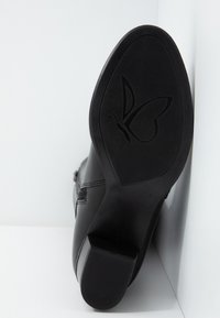 Zapato de cuero negro con una suela de goma texturizada que presenta un logo de mariposa, colocado en un ángulo sobre un fondo blanco.