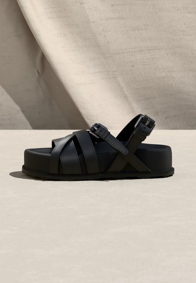 Μαύρο πλατφόρμα σανδάλι με σταυρωτούς ιμάντες, ρυθμιζόμενες αγκράφες και υφασμένο πέλμα σε φόν από ελαφρύ ύφασμα και σκυρόδεμα.