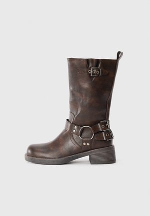 Bota de cuero marrón hasta la rodilla con punta cuadrada, una correa con una hebilla circular, herrajes en tono plateado y un tacón cuadrado texturizado.