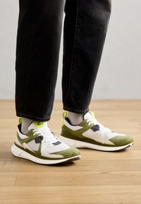 Weiße und grüne Sportsneaker mit Mesh- und Wildledertexturen, schwarzen Akzenten und einer abgerundeten Sohle. Tragen mit schwarzen Jeans und grauen Socken.