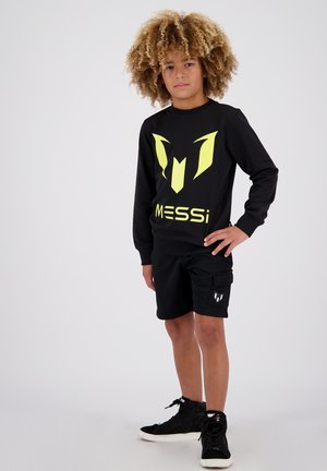 Fekete pulcsi neon sárga logóval és részletekkel, fekete rövidnadrággal, amely oldalsó zsebekkel rendelkezik, és fekete magas szárú sportcipőkkel viselve.