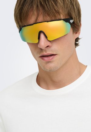 Jeune homme portant une chemise blanche et de grandes lunettes de sport jaunes réfléchissantes couvrant ses yeux et le haut du nez.