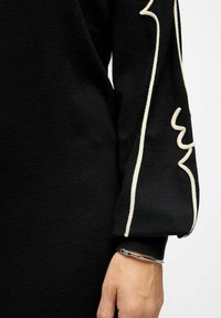 Pullover nero a maglia con un design bianco in evidenza sulla manica. Texture liscia con polsini aderenti; un bracciale d'argento al polso aggiunge contrasto.