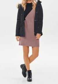 Parka nera con rifiniture in pelliccia sintetica, maglione blu navy e vestito in maglia rosa. Abbinato a stivali neri alla caviglia con lacci.