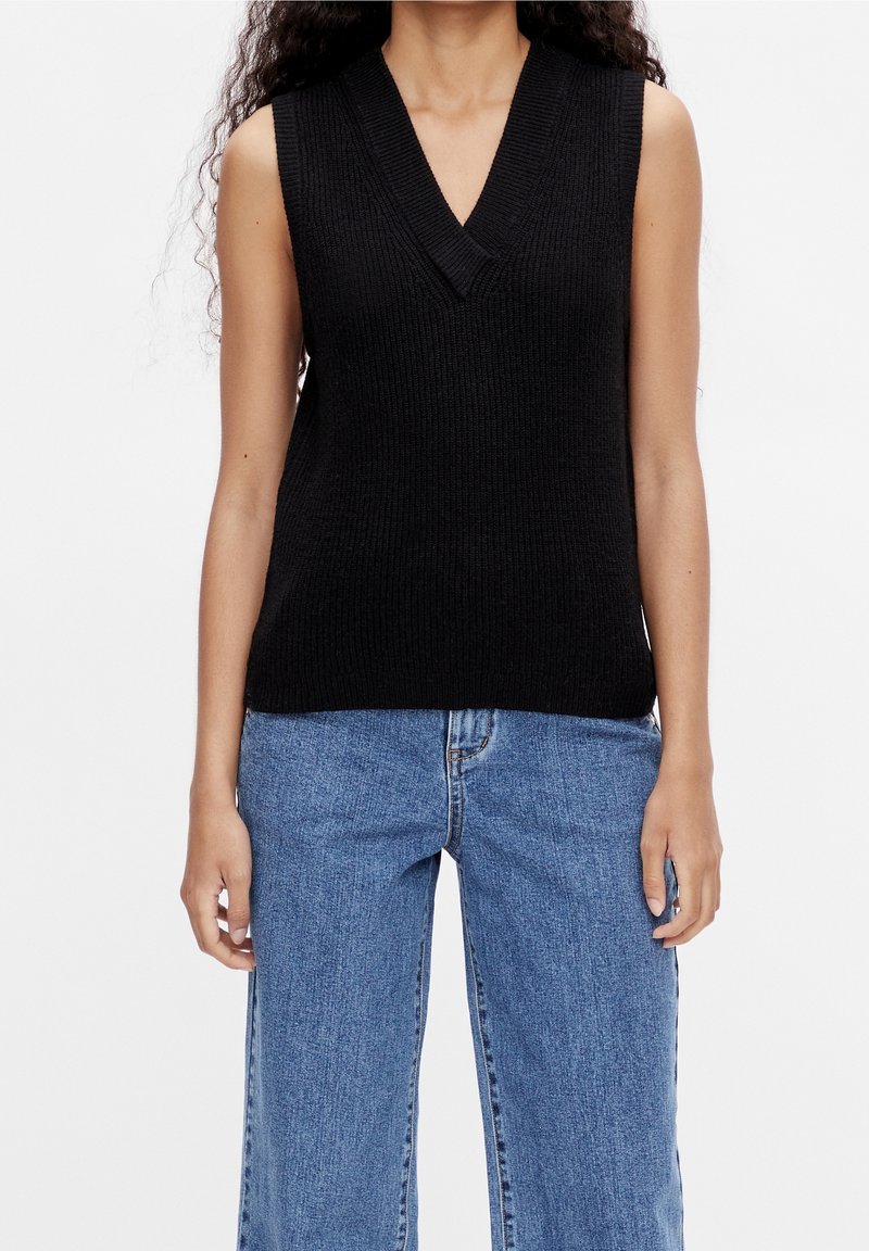 Femme portant un débardeur en maille noire sans manches avec un col en V et un jean bleu taille haute, debout devant un fond uni.