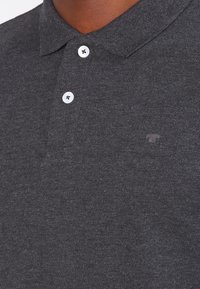 Mörkgrå pikétröja i strukturerad tyg, med en klassisk krage, tre vita knappar och en liten logotyp på bröstet.