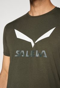 Oliven grønn t-skjorte med korte ermer, med et hvitt grafisk logo som ligner vinger og "SALEWA" i en reflekterende skrift på forsiden.