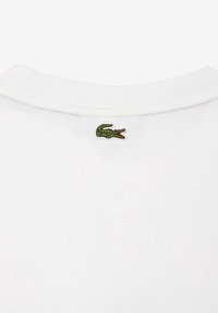 Camiseta blanca de algodón con un pequeño logo de cocodrilo verde bordado en la parte posterior del cuello. Textura suave, diseño de cuello redondo.