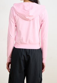 Bakåtsikt av en person som bär en ljusrosa kortärmad hoodie och lösa svarta cargobyxor mot en neutral bakgrund.