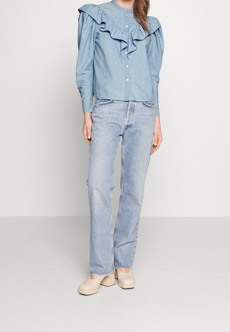 Femme portant un chemisier bleu clair à volants avec des épaules bouffantes, un jean droit délavé clair, et des chaussures beiges à talons épais, se tenant devant un fond uni.