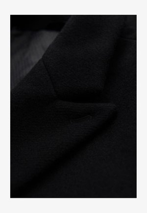 Tessuto di un cappotto di lana nera con una superficie liscia, caratterizzato da un colletto strutturato e un piccolo dettaglio di tasca discreta sulla patta.