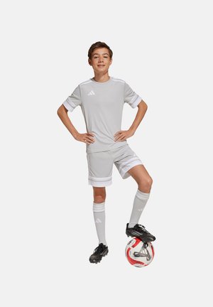 Een jeugdige in een lichtgrijze Adidas voetbaluitrusting, met korte mouwen en strepen, staat op een rood-witte voetbal en draagt zwarte voetbalschoenen.